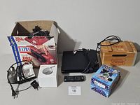 Full lot view showing all items boxed and unboxed on table: Dirt Devil Ultra vacuum in box, LG DVD player with remote, M&M mini plush radio in box, Coca Cola clock radio, wireless Bluetooth speaker, Proctor Silex iron