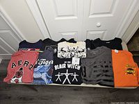 Top view of nine men's graphic t-shirts laid flat, showing different colors and large visible graphic designs like Aero, Blair Witch, and Reese's.