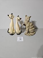 Three ceramic cat figurines displayed on white surface against white background, showing two cream elongated cats on left and one grey striped cat on right