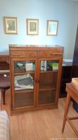 Front view of the wooden glass door display cabinet with some contents inside. Shows two glass doors, two drawers above, and flat top surface.