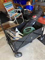 Wide angle showing black folding cart filled with watering cans, bug swatters, and folded bag chair with multicolored stripes.