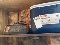 Motomaster battery charger and Coleman cooler on a shelf with insulation behind