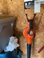 Full view of the handheld cordless blower hanging on wood wall with trash bins in background.