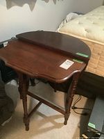 Both demi lune tables side by side, showing the smaller gate leg style table with turned legs and the Bombay style table with curved legs and brass knobs, against a background of a mattress and electronics.