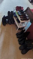 Overview of footwear arranged around and on a small white two-level plastic shoe rack on carpet floor. Includes various boots, slippers, sandals, shoes in predominantly black and brown shades.