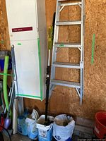 Photo shows 6 foot aluminum stepladder hanging on wooden wall next to a white storage cabinet and a Radley cordless pole saw leaning in front of bags of driveway salt.
