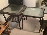 Photo showing two outdoor tables together, illustrating size difference and materials: resin wicker and coated metal, each with glass tops.