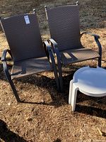 Two brown metal framed patio chairs with woven synthetic seats and backs and a small white plastic round table taken outside on dry grass.
