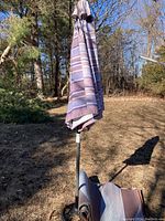 Patio umbrella with striped purple and white fabric canopy, metal stand, and rolled matching matting on ground beside it