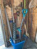 Photo showing assorted shovels, red snow rake, the blue tub containing tools, and a fishing or gardening net hanging on the wall.