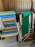 Photo showing three folding chairs (two green canvas, one multicolored striped canvas) in front of a large beige Rubbermaid storage container