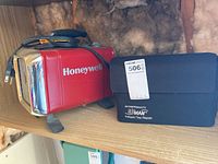 Photo of Honeywell portable heater in red with chrome grille and black base next to AirMan tire repair kit on wood shelf.