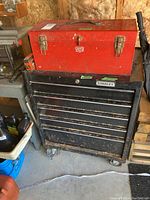 Red metal toolbox atop black Stanley tool chest with drawers and wheels in a workshop setting.