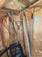 Photo of five garden tools hanging against wooden wall including a bow saw, two garden rakes, a shovel, and a hoe.