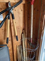 Two long-handled hedge shears with wooden handles and black blades, a wire plant staking ring leaning against wall, and a red-edged garden edger.