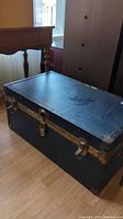 Black vintage steamer trunk with brass locking latches and scuffs on top surface shown closed.