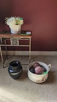 Photo showing small metal frame table with wooden top, black ceramic vase on floor, and cream ceramic pot with decorative balls next to it