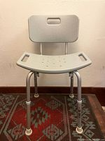 Front view of Drive shower/bath chair with backrest, adjustable legs, and rubber tips on each leg. Mounted on decorative rug against wall.