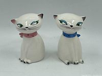 Front view of the pair of white ceramic cat-shaped salt and pepper shakers with pink and blue collars on plain background.