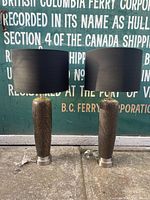 Pair of bedside table lamps with textured brown bases and tall black lampshades with gold interiors shown front view on pavement against a green backdrop with white text.