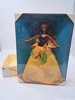 Barbie doll in sunflower themed dress inside original display packaging with blue backdrop. Doll has curly hair, yellow and orange dress resembling sunflower petals and leaves.