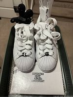 Front view of white Adidas Superstar 35th Anniversary shoes with thick white laces and additional black and white laces attached, sitting on the shoe box showing the 35th Anniversary text.