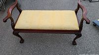 Front view of antique style storage bench with yellow upholstered seat and carved wooden armrests and legs.