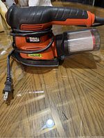 Single power sander with clear dust collection container and wrapped power cord resting on wooden surface.