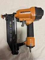 Orange and black Bostitch pneumatic nailer with 16 gauge capacity and textured handle laid on white surface, showing side profile of the tool.