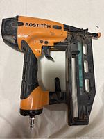 Side view of a used Bostitch pneumatic nailer in orange and black with visible wear and attached air hose connector.
