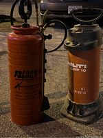 Two concrete sprayers side by side; red Form & Build sprayer with black pump handle and hose on the left and red Hilti DWP 10 sprayer with black top and handle on the right.