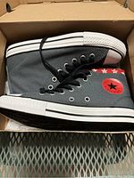 Side view of grey Converse Chuck Taylor All Star high top sneakers showing the red logo patch and black shoelaces, inside original cardboard box.