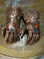 Pair of bronze-colored toy gloves modeled after the Infinity Gauntlet featuring six colored gems, laid flat on a wooden surface.
