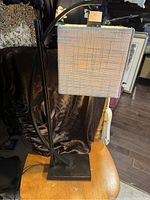 Photo of a black metal table lamp with an arched arm holding a rectangular, beige checkered fabric lampshade sitting on a wooden table.