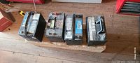 Four used marine batteries lined up on wooden floor, seven visible handles, dusty condition