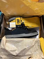 Photo showing the right side profile of one black CAT safety shoe with grey sole inside original box with yellow tissue paper and CAT branding tag attached to the shoe.