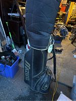 Photo of black Mizuno branded golf bag standing upright on floor in a cluttered space.