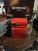Red Nespresso Inissia espresso coffee machine displayed with original box and instruction manual on round wooden table.