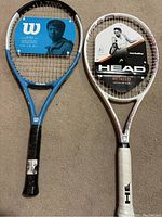 Photo showing two brand new tennis rackets side by side on a carpet. The left racket is Wilson Power Tilt in blue and white with a black grip. The right racket is Head Metallix in white and metallic with a white grip. Both have retail labeling attached.