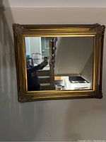 Square gold framed wall mirror with decorative corners and beaded inner molding, reflecting indoor room scene.