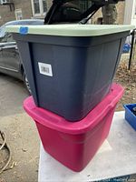 Two stacked plastic storage totes with lids, one dark gray with light gray lid, one maroon with maroon lid, shown outdoors on pavement near vehicles and fence.