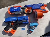 Three Nerf blasters laid on a surface: one large Nerf Elite blaster with an orange drum magazine, a black and blue Nerf Maverick Rev-6, and a small blue and orange Fortnite toy blaster with darts.