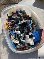Plastic container filled with a variety of LEGO bricks and plates in multiple colors and shapes, including some with wheels and printed elements.