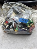 Plastic bag containing a variety of LEGO bricks and small elements including some flat tiles and specialized pieces, visible in multiple colors.