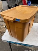 Photo of a large yellow Rubbermaid Roughneck storage tote with lid sitting on a table, showing the lid and part of the container body with reinforced sides and handles.