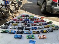 Wide view of all Hot Wheels toy cars on a white table outdoors showing variety and colors.