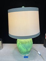 Full view of the Art Deco parrot glass table lamp with shade, showing the lamp lit and the beige fabric shade with blue trim.