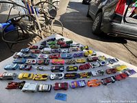 Photo showing approximately 60 Hot Wheels toy cars laid out on a white surface outdoors, displaying a wide variety of colors and models.