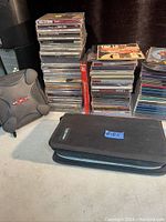 Wide shot of multiple stacks of used jewel case CDs including a small black carrying case and a gray speaker on a table.