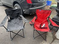 Two folding camping chairs, one black and one red, shown outdoors on pavement near a vehicle.